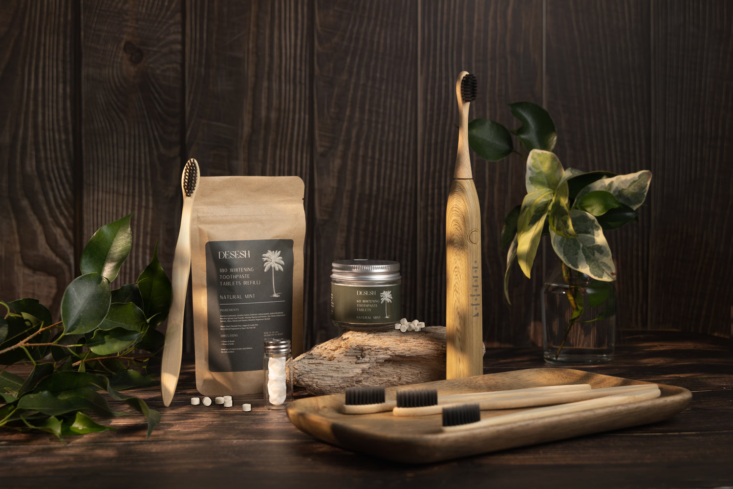 This image shows a glass jar of toothpaste tablets, a biodegradable toothpaste tablet pouch, biodegradable dental floss, and bamboo electric and manual toothbrushes. The items are on a wood surface next to plants. These are a part of Desesh's range of zero waste, plastic free, eco friendly, more sustainable personal care and everyday essential products.