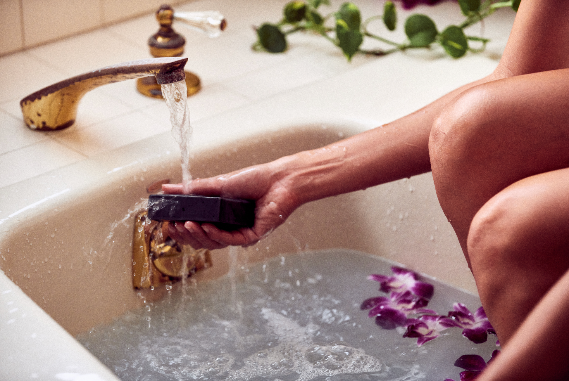 This photo shows the Desesh premium quality US-made small batch body hand and face soap bar with plastic free biodegradable packaging. In the image a women is in the bath running water over the bar before lathering and washing her hands