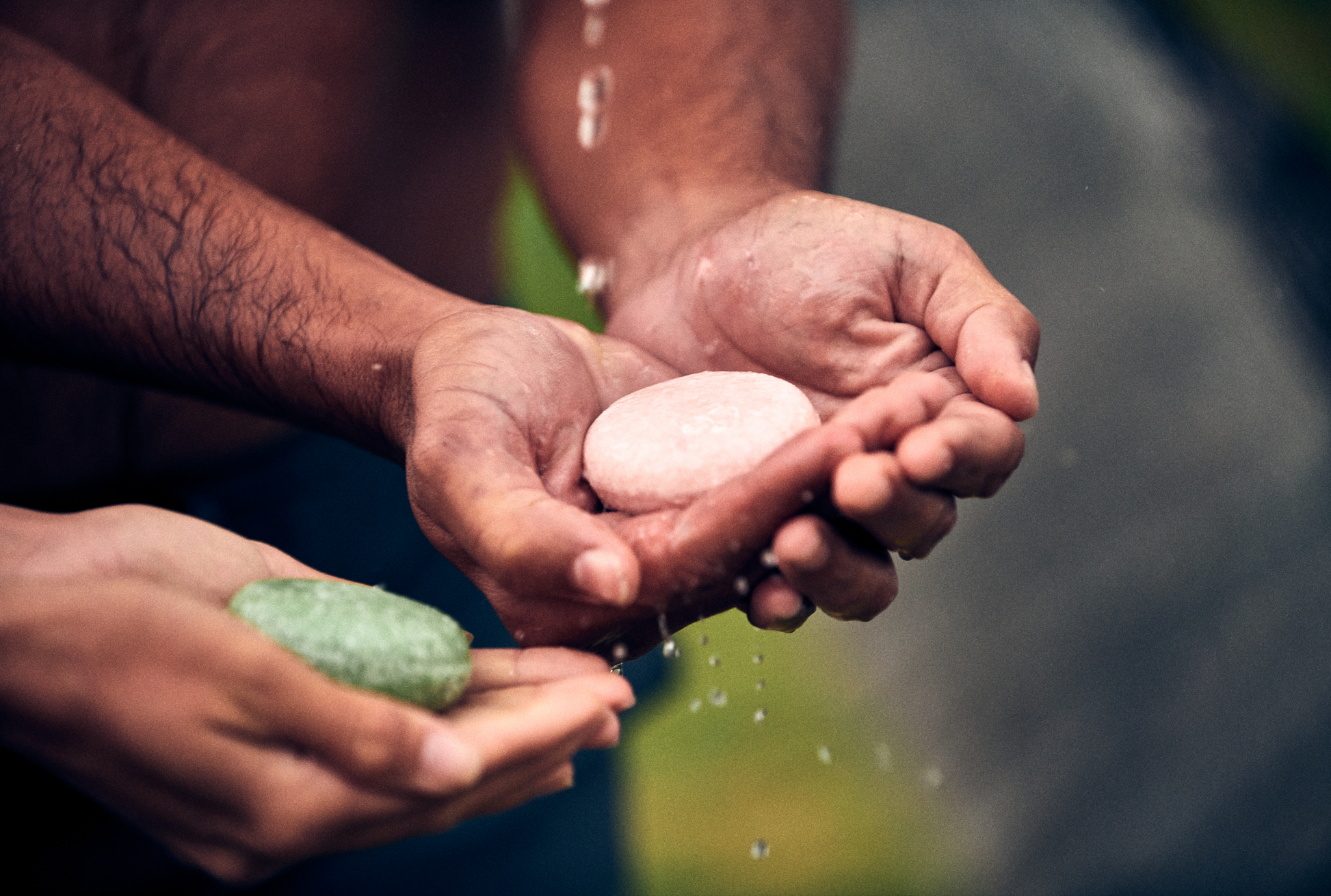 This image shows salon quality Argan Oil Shampoo bars with plastic free biodegradable packaging for a more sustainable eco friendly zero waste hair care routine. In the image two people are holding a Tea Tree and Mint solid shampoo bar and a Lavender and Lime solid shampoo bar and are applying water to them before washing their hair.