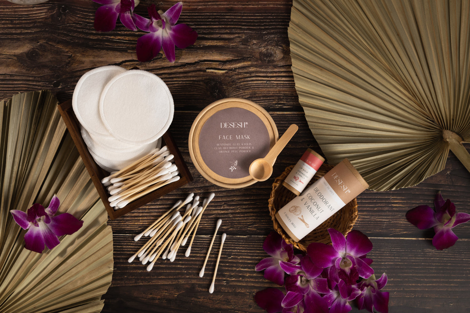 This image shows a range of plastic free skincare products including natural aluminum free deodorant, natural lip balm, face mask powder, reusable cotton buds and rounds. The products are on a wood surface next to orchids and dried palm leaves. These are a part of Desesh's range of zero waste, plastic free, eco friendly, more sustainable personal care and everyday essential products.