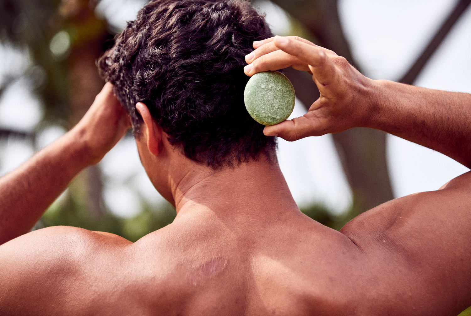 This image shows a shirtless man in an outdoor setting holding a Tea Tree and Mint Solid Argan Oil Shampoo Bar next to his head. These are a part of Desesh's range of zero waste, plastic free, eco friendly, more sustainable personal care and everyday essential products. 