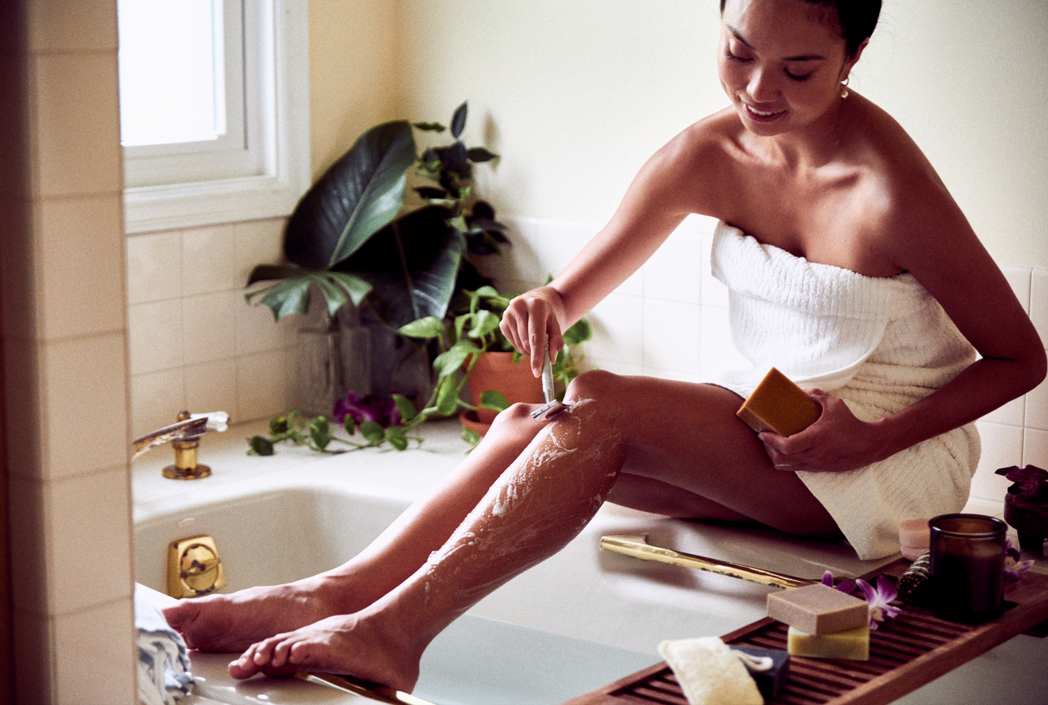 This image shows a woman sitting on a bathtub for her low waste plastic free shaving routine. She is using a metal safety razor and a solid shave soap bar. These are a part of Desesh's range of zero waste, plastic free, eco friendly, more sustainable personal care and everyday essential products.