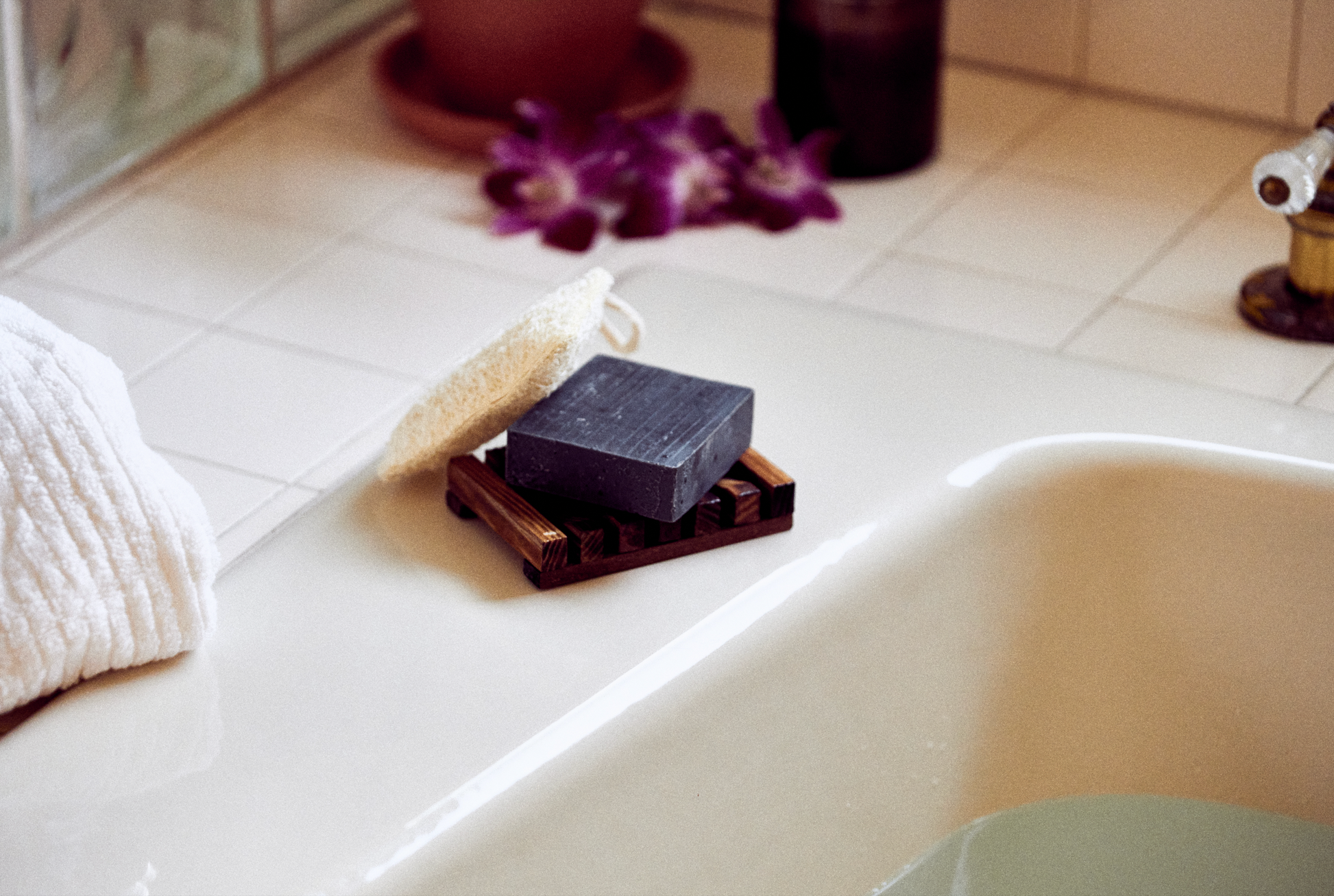 This image shows a dark stained wood soap dish to help keep soap dry. It is rectangular in shape. It is designed for a more eco friendly zero waste sustainable lifestyle and routine. This image shows a bar of natural soap on top of the soap rack and next to a natural loofah exfoliating pad for skin care