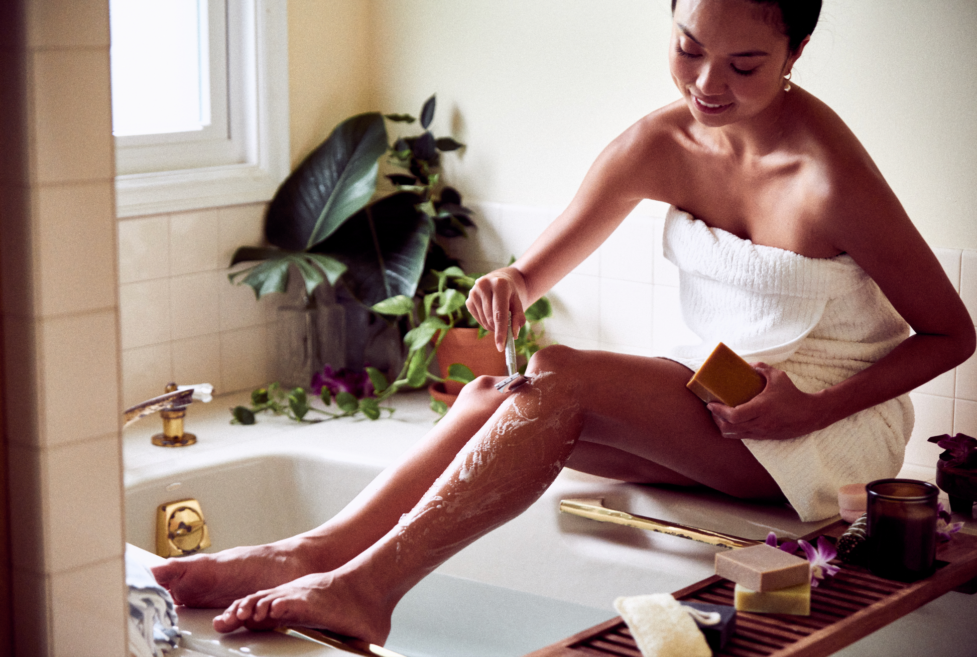 This image shows the Desesh premium quality solid body and face shave soap bar as an alternative to cans of shave foam or cream. This is a more eco friendly sustainable zero waste option and the shave bars come in biodegradable packaging. Shown here is a woman using a metal safety razor to shave her legs in the bath while using a solid shave soap bar