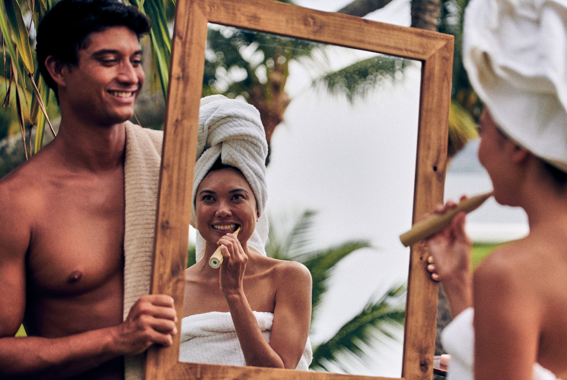 This image shows a woman brushing her teeth with a sonic bamboo electric toothbrush and US-made fluoride free mint toothpaste tablets while looking into a mirror that a shirtless man is holding